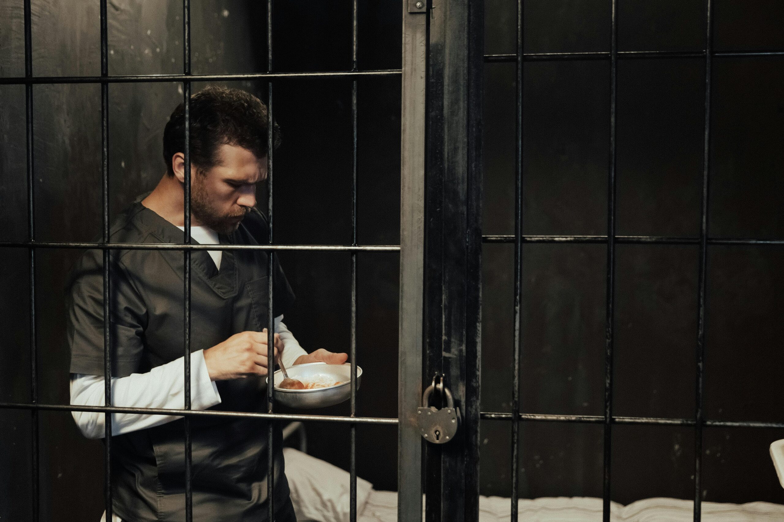 A male prisoner stands in a jail cell, eating a meal from a bowl.