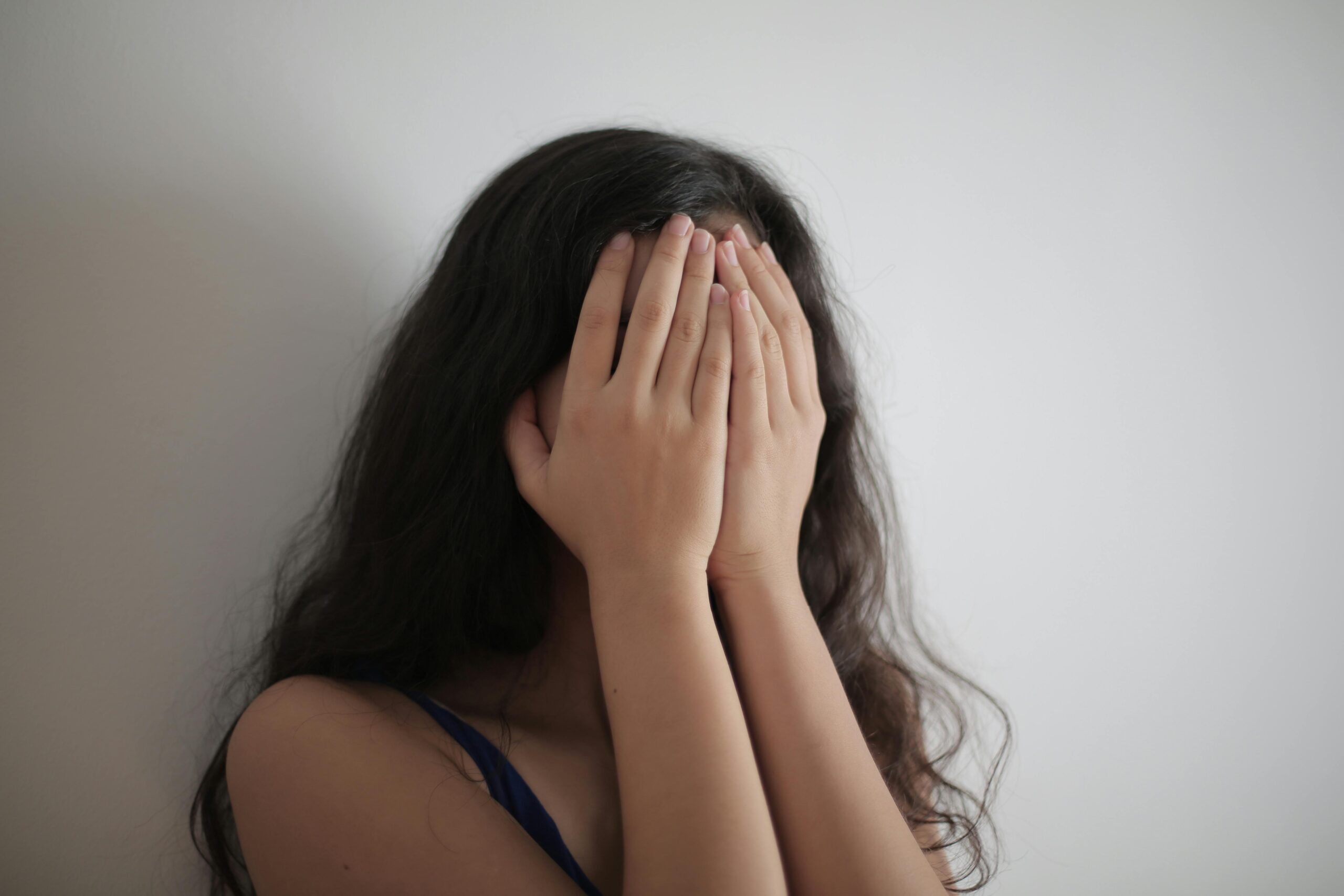 An adult woman with long brunette hair covers her face with her hands against a white wall, creating a minimalist portrait.