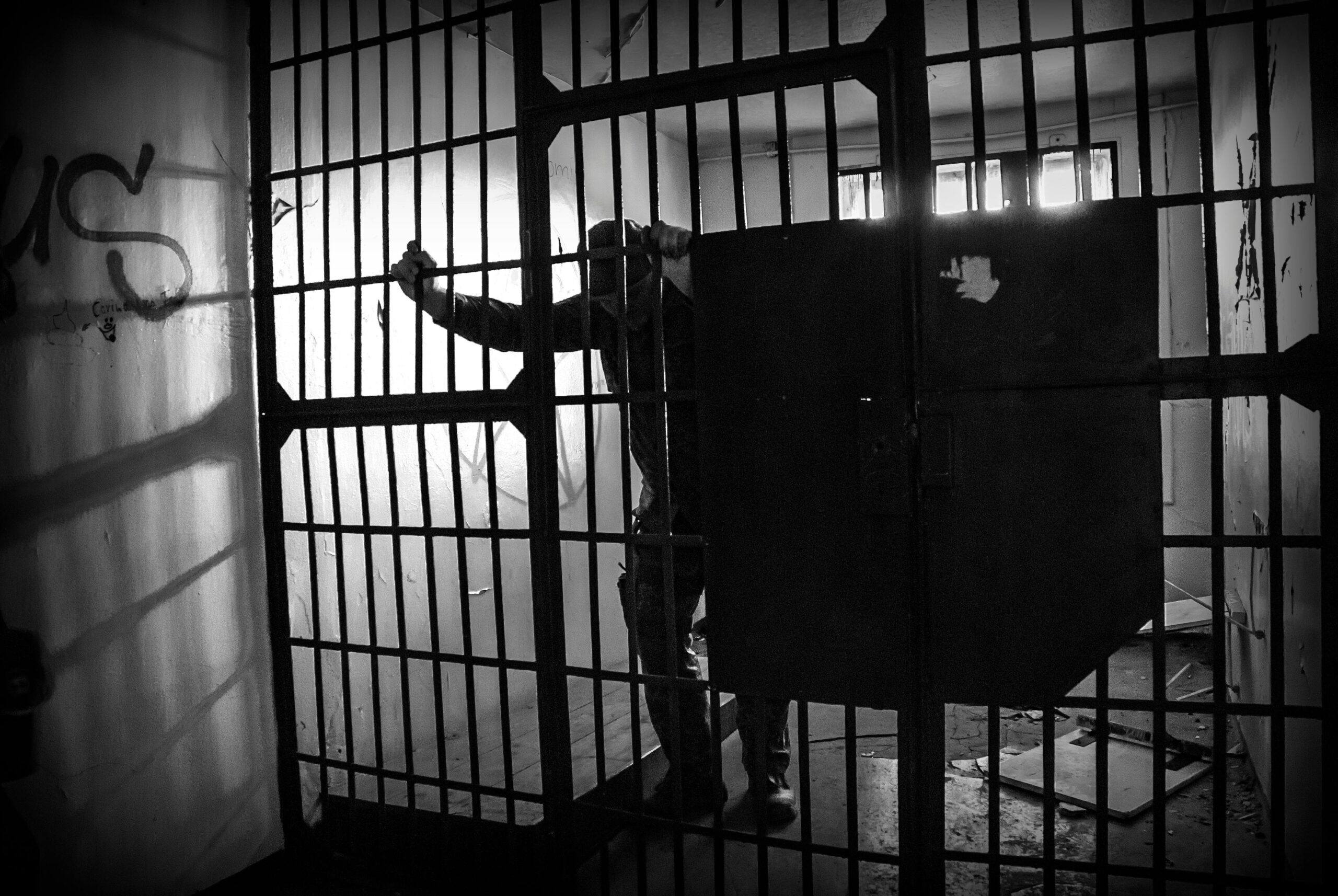 Black and white image of a lone prisoner behind bars inside a dimly lit cell.