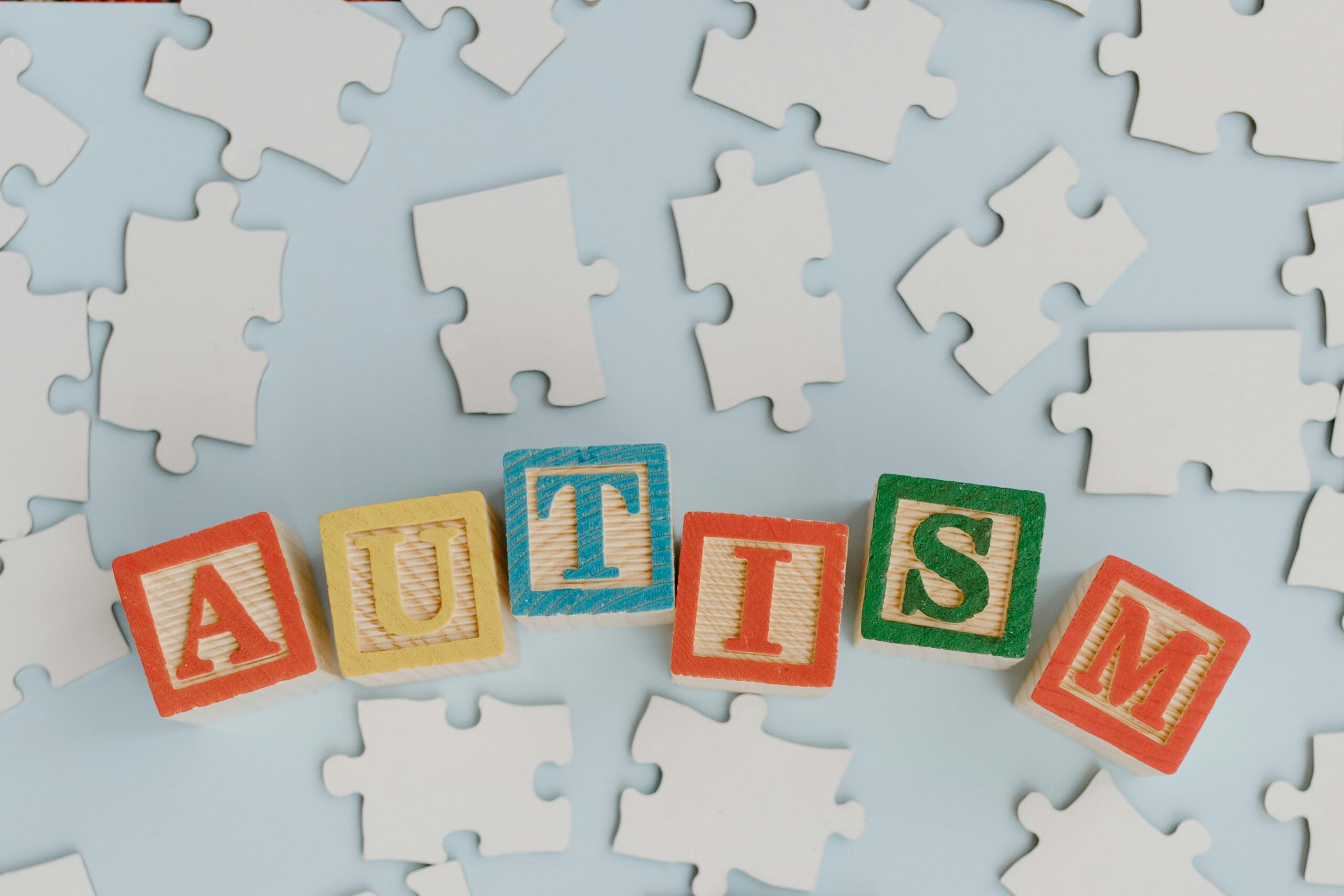Colorful blocks spelling 'autism' amidst scattered puzzle pieces on a light blue background, symbolizing autism awareness.