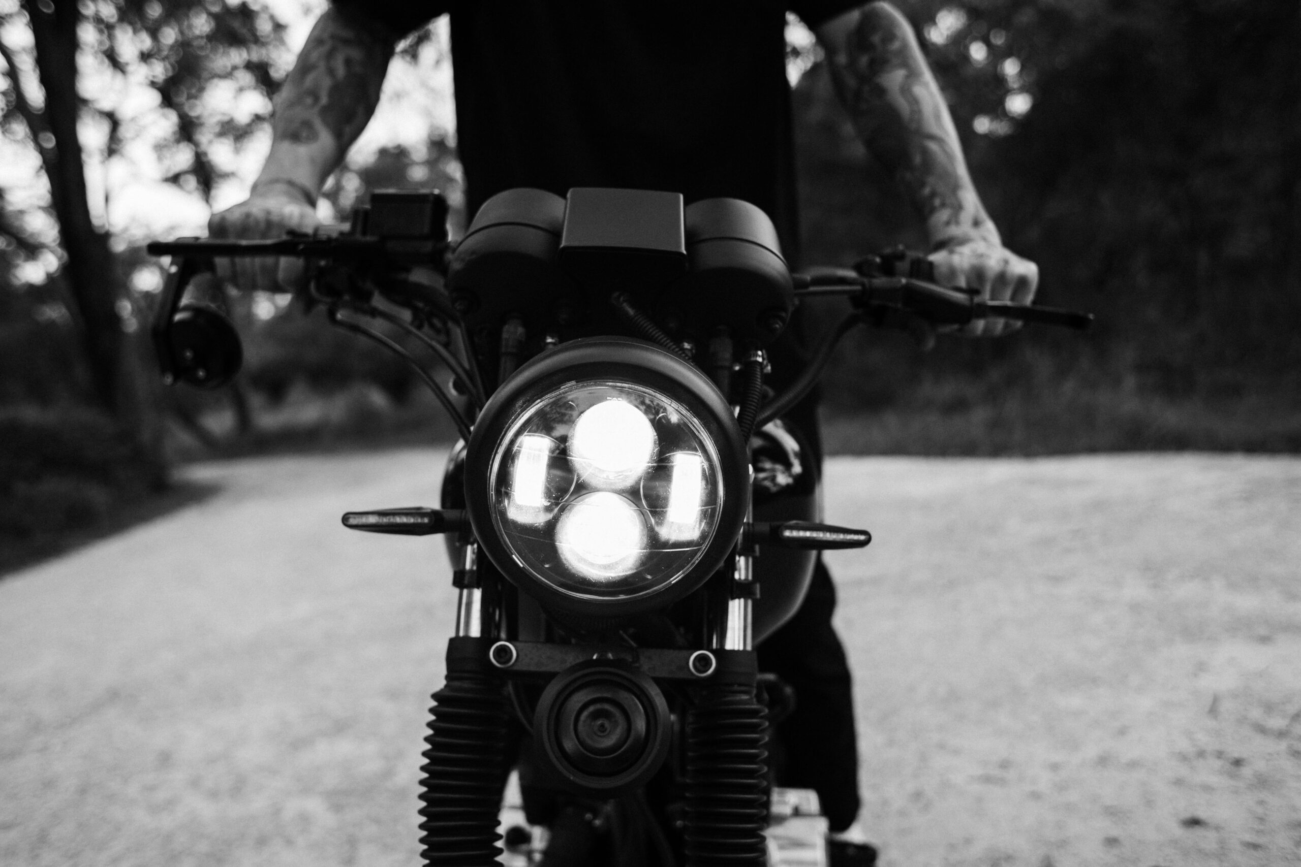 A tattooed person rides a motorcycle down a rural road in a dynamic black and white shot.