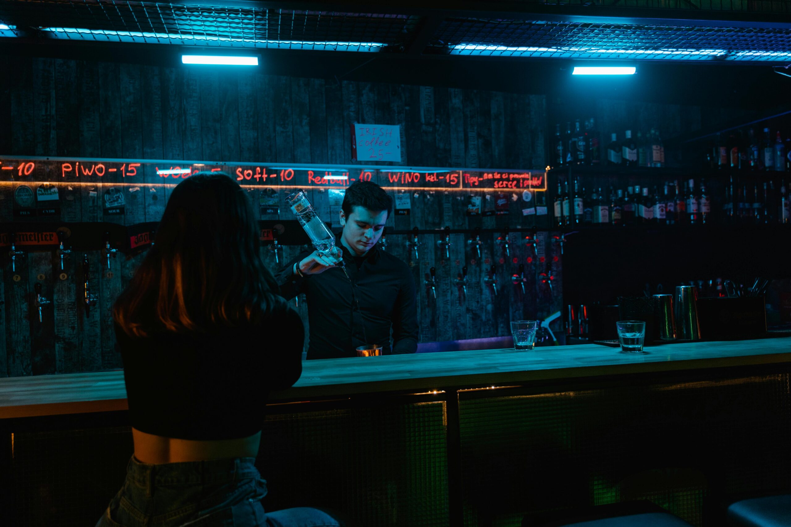 A bartender pours a drink under neon lights in a moody bar setting, with a patron seated at the counter.