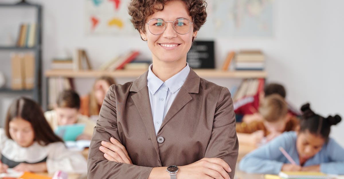 Smiling teacher with glasses standing with arms crossed in a classroom, students learning.