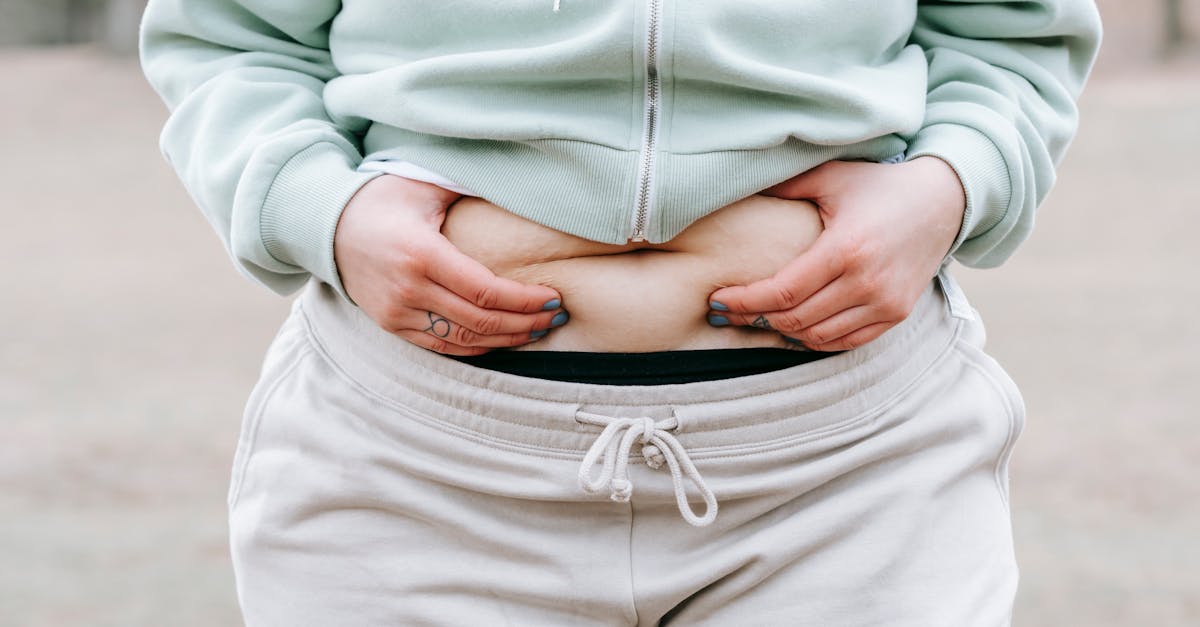 Crop anonymous plus sized female in casual hoodie touching and showing fat on belly while standing in park